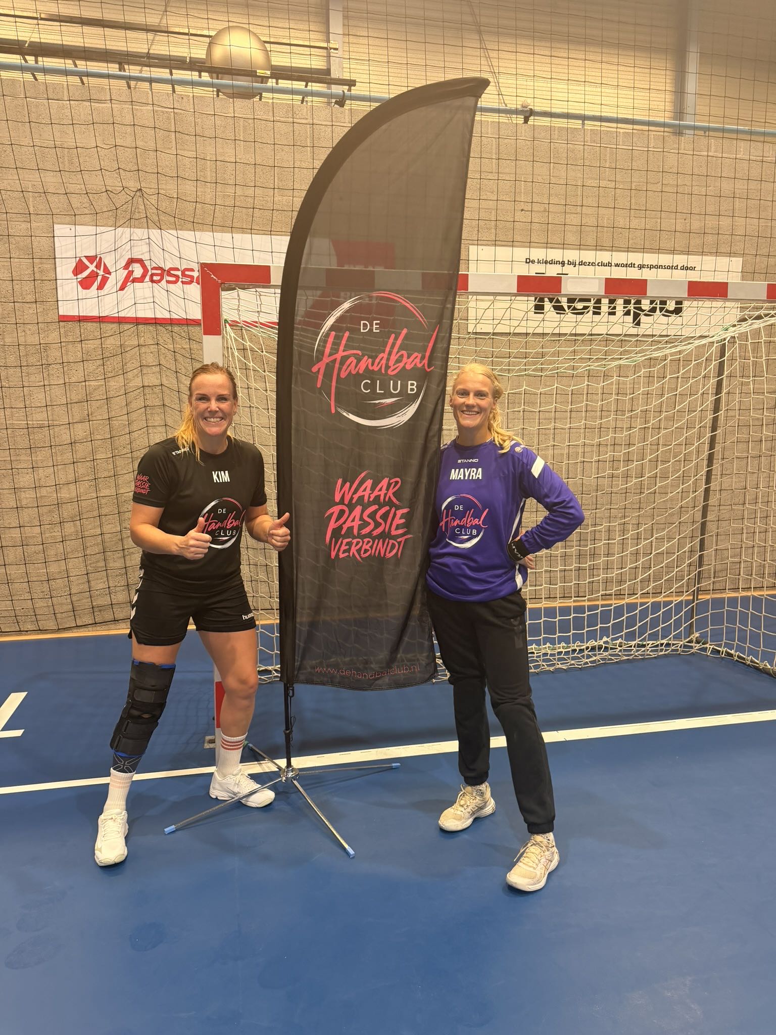 Kim in het zwarte speelshirt en Mayra in het paarse keepersshirt, naast de beachflag van de Handbalclub met de tekst Waar passie verbindt