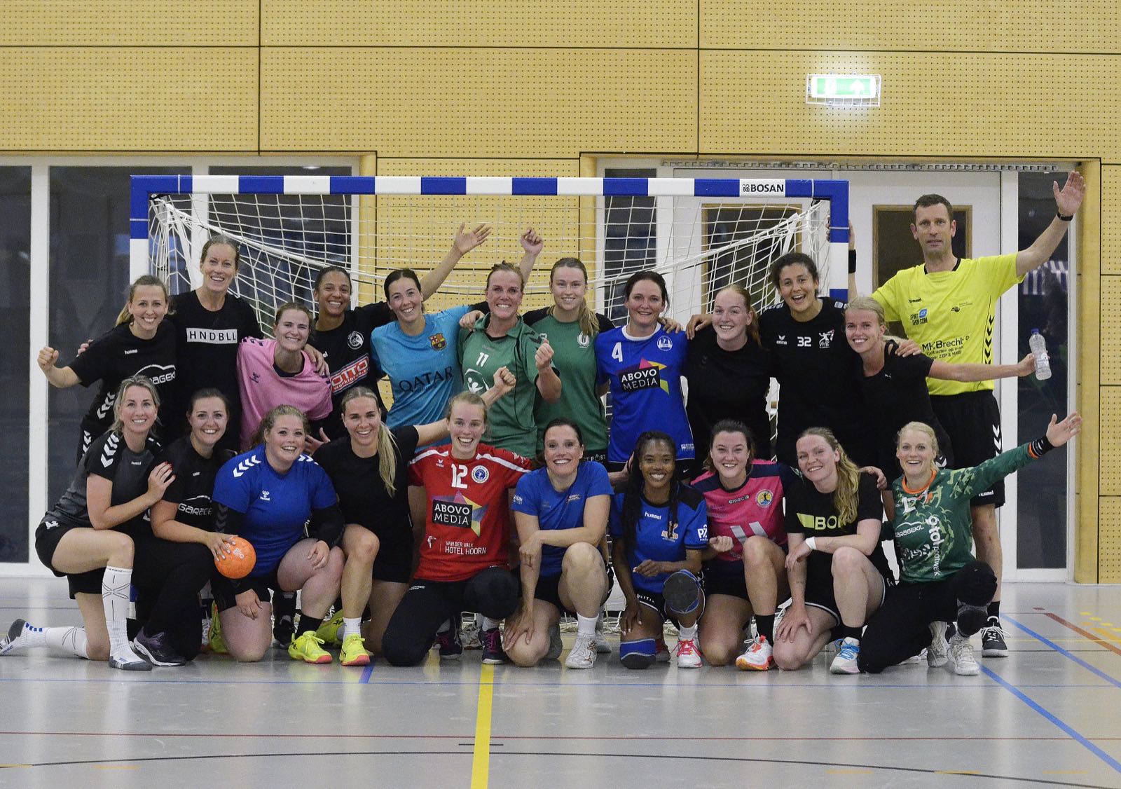 Groepsfoto van ruim twintig speelsters en de scheidsrechter voor het doel in de sporthal, na de tweede Match Night van de Handbalclub op 5 juni